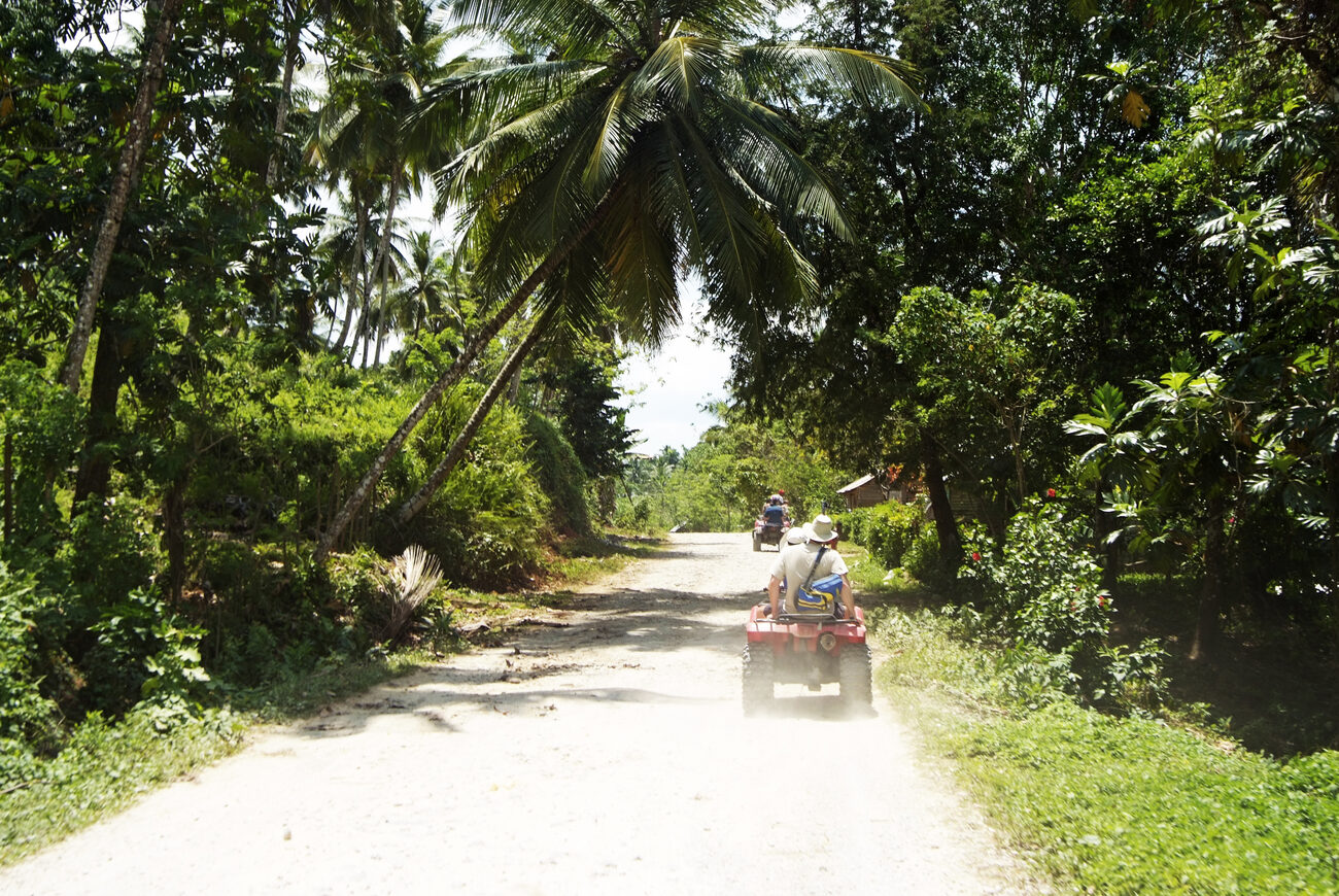 Two ATVs drive along a gravel road through the jungle.