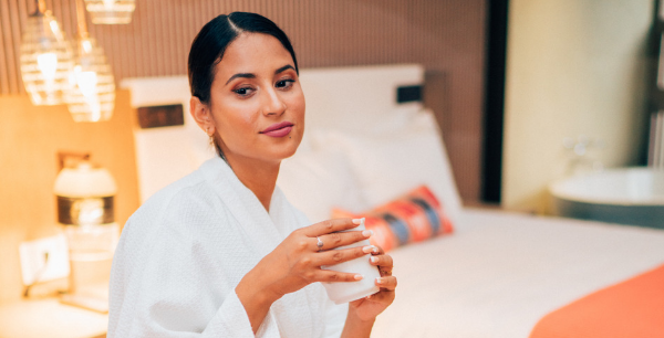 A woman in a white robe sits on the hotel bed with a mug in her hands.