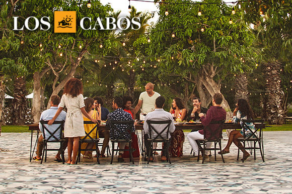 A group of adults are gathered at a table outside beneath dangling lights.